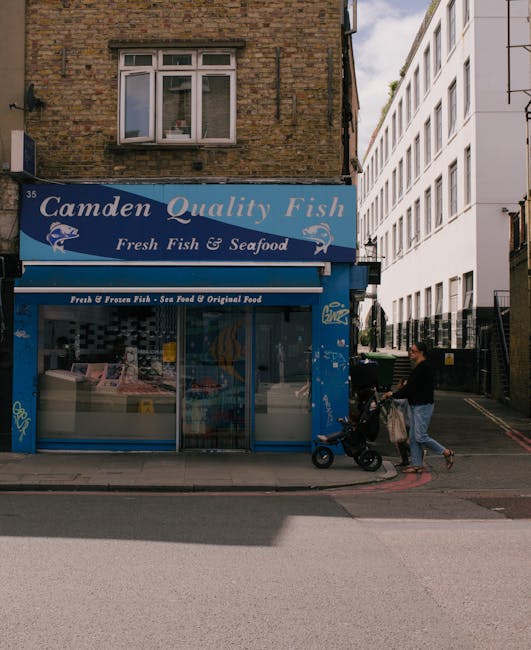 A street-level view of a blue storefront sign reading 'Camden Quality Fish' with additional signage indicating 'Fresh Fish & Seafood' and 'Fresh & Frozen Fish - Sea Food & Original Food.' The shop has