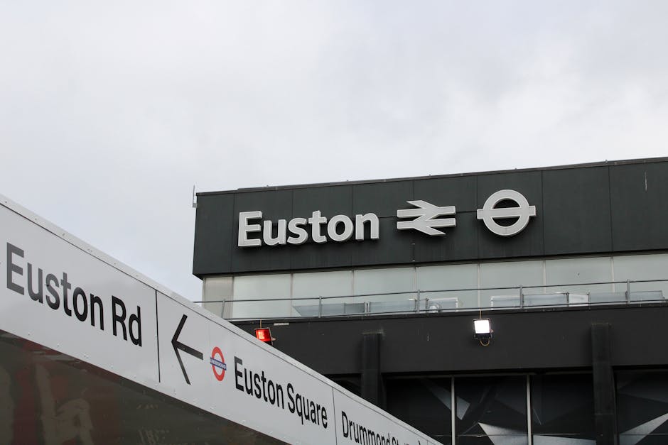 A close-up view of a brick building with arched windows showing yellowish-brown bricks and dark window frames. A rectangular sign with the red and white logo of a British railway company, reading 'Bic